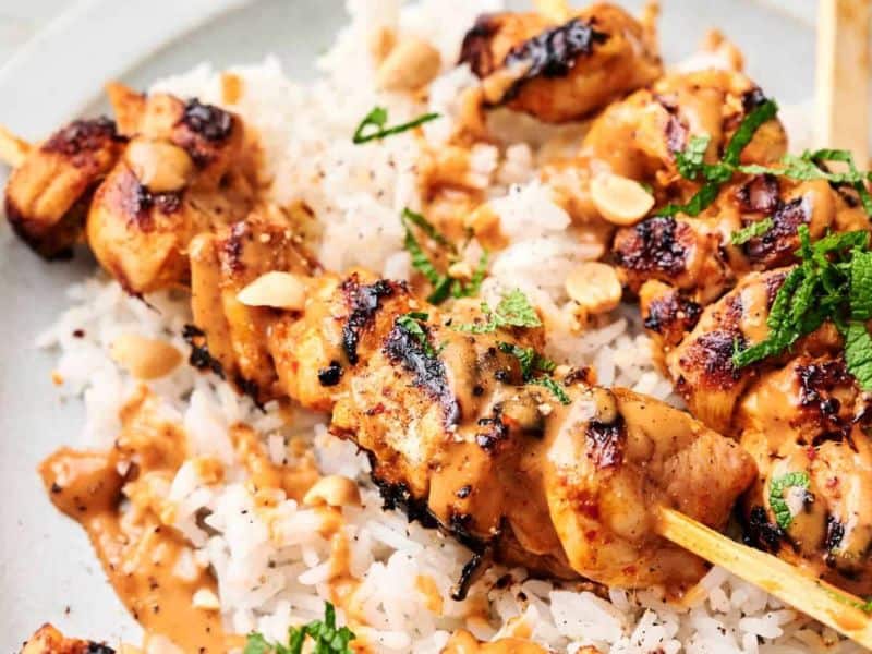 Thai chicken skewers topped with a creamy sauce and garnished with chopped herbs and peanuts, served over a bed of white rice.