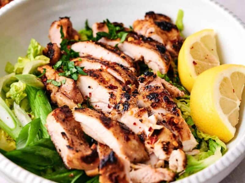 Grilled chicken breast slices served over a bed of greens with lemon wedges on the side.