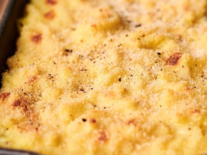 Close-up of a creamy, golden-brown macaroni and cheese dish with a crunchy breadcrumb topping reminiscent of comforting funeral potatoes. A spoon is partially submerged in the cheese sauce.