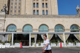 A woman wearing a red jumper and sunglasses, as well as a little bag on her right shoulder, talks on the phone in front of an exterior view of the Central Bank of the Russian Federation building on top of which the Russian flag can be seen
