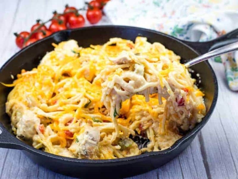 A hand holding a fork with a serving of chicken spaghetti from a cast-iron skillet.