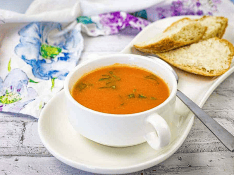 A bowl of old-fashioned tomato soup garnished with herbs, served with slices of crusty bread on a white plate.