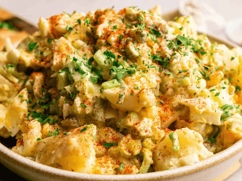 A bowl of potato salad topped with chopped herbs and a sprinkle of paprika, with visible chunks of potatoes and vegetables.