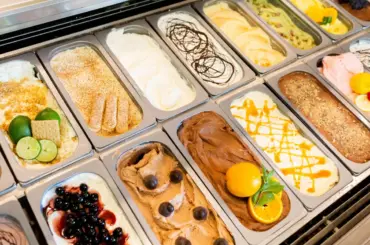 Assorted gelato flavors displayed in a metal case, including chocolate, fruit, and nut varieties, some topped with fruit slices, sauces, or cookies—each scoop inviting you to experience The Sweet Life.