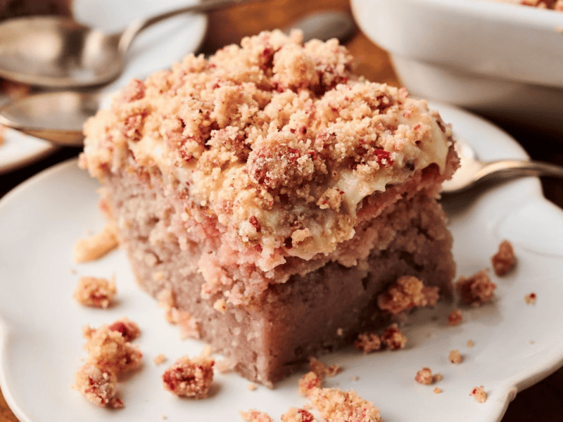A slice of crumb-topped strawberry coffee cake sits on a white plate with a spoon beside it; additional cake and plates are in the background.