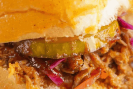 Close-up of a pulled pork sandwich with pickles and red cabbage on a bun, held in hand, with chips in the background.