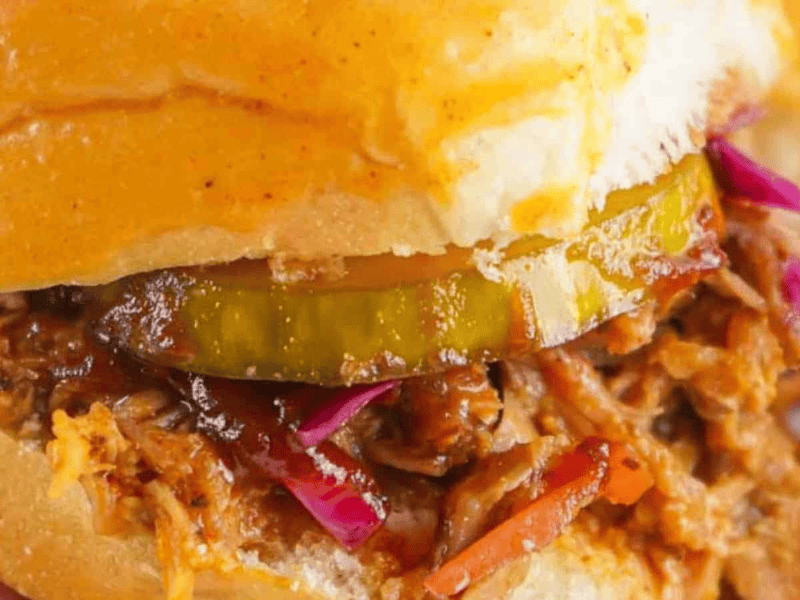 Close-up of a pulled pork sandwich with pickles and red cabbage on a bun, held in hand, with chips in the background.
