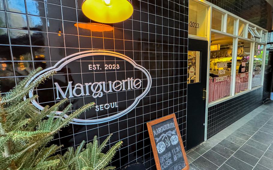 A close-up view of a restaurant facade, with a door next to a black stone wall with a white logo reading “Est. 2023 Marguerite Seoul.”
