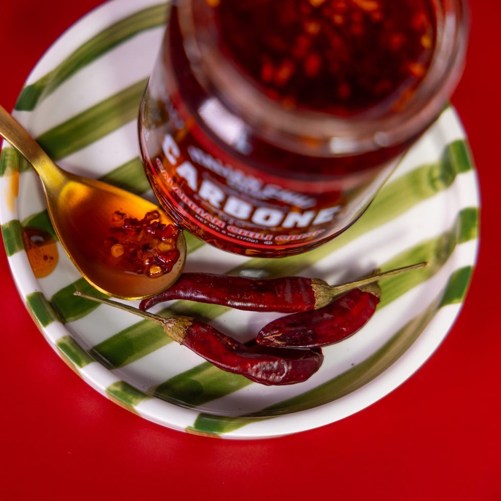 Golden spoon with chili flakes beside a jar of chili oil and dried red peppers on a striped plate against a red background. Golden spoon with chili flakes beside a jar of chili oil and dried red peppers on a striped plate against a red background.