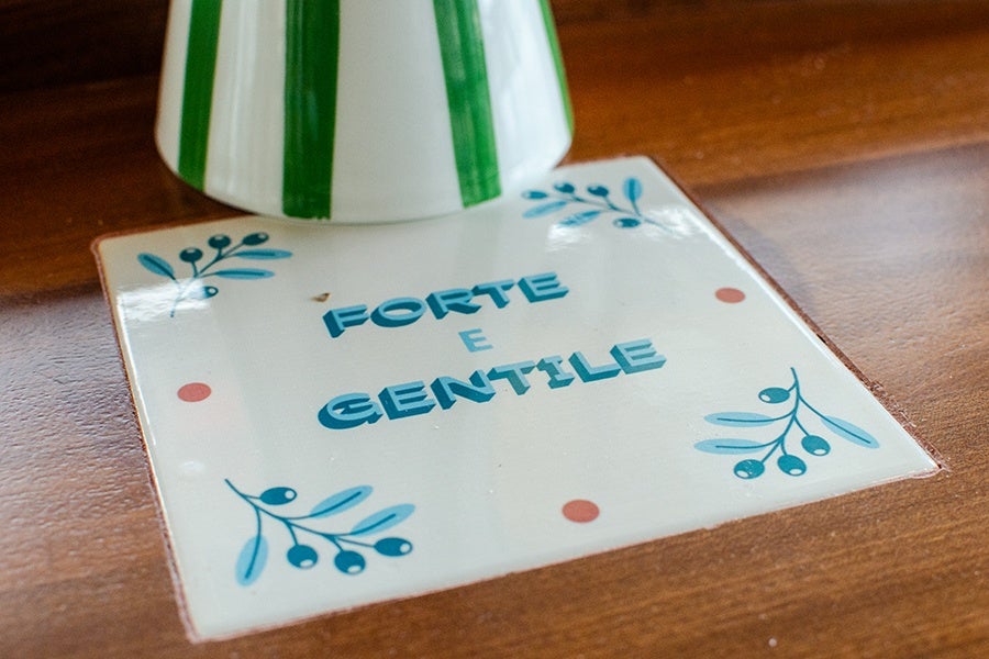 A white ceramic tile set into a wooden surface, decorated with blue leaves and berries in the corners and four small red dots near the edges. The tile has the Italian words 
