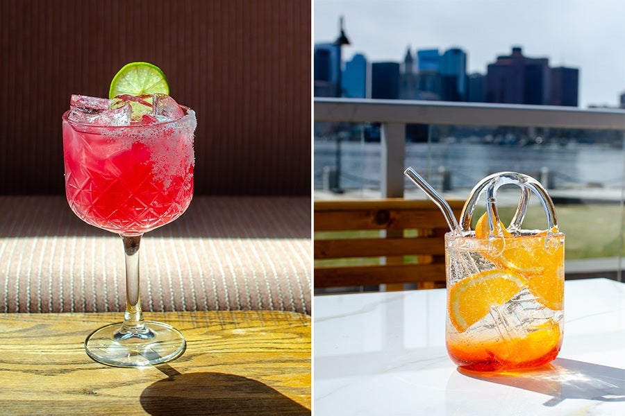 A red cocktail in a decorative glass with a salted rim, ice cubes, and a lime wheel garnish on the left. On the right, a clear glass with a handle containing a sparkling orange drink with ice, orange slices, and a metal straw, set on a white table with a waterfront cityscape in the background.