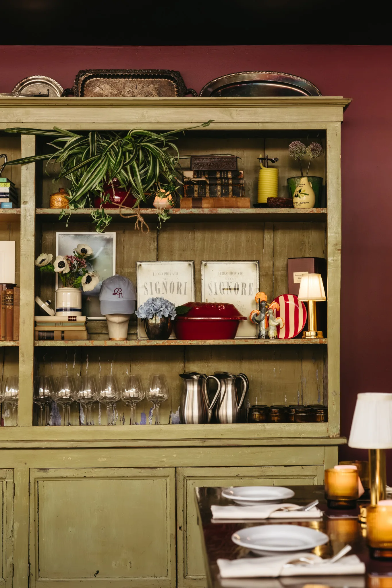 Green wooden cabinet with shelves displaying dishes and decorative items, and partially visible dining table.