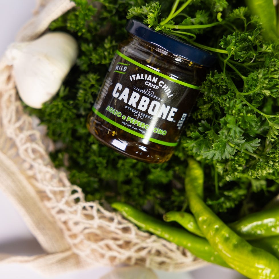 Jar of Carbone Italian chili crisp surrounded by fresh parsley, garlic, and green peppers in a natural bag. Jar of Carbone Italian chili crisp surrounded by fresh parsley, garlic, and green peppers in a natural bag.