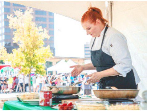 A person in an apron preparing Italian food at an outdoor cooking station with festival attendees visible in the background.