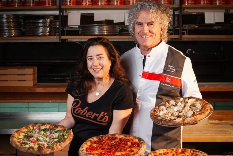 This photo shows Dana Hokin and Robert Garvey, each holding pizzas, with more pizzas on the table in front of them. Robert is wearing his 50 Top Pizza chef coat.
