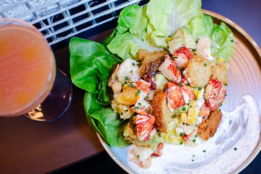 Overhead view of a salad with chunks of lobster, big croutons, bibb lettuce, and a swoosh of herby white sauce, next to a cocktail.